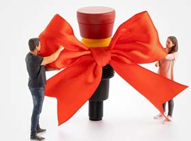 Bild zeigt Miniaturfiguren 2er Mitarbeiter die eine rote Schleife an einem riesigen Befehlsgerät befestigen